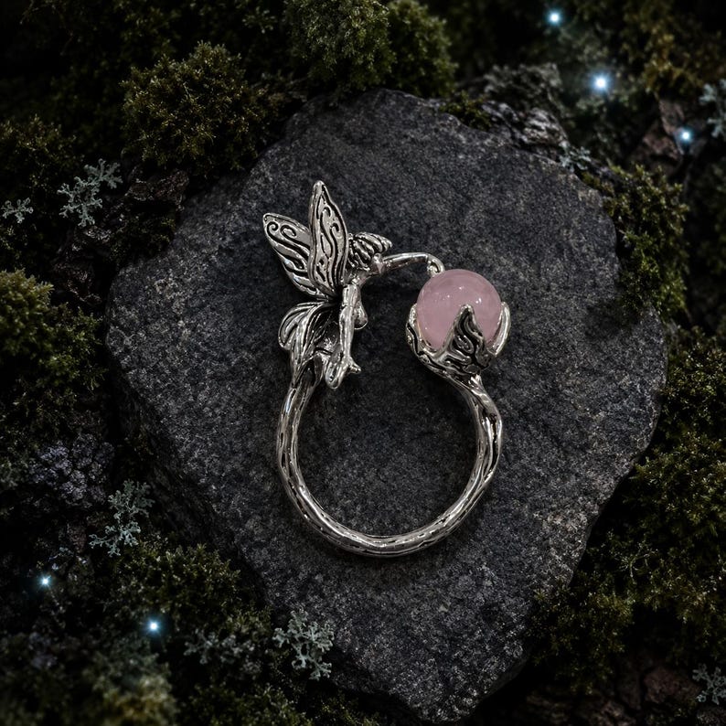 May include: A silver-toned ring featuring a fairy figure with detailed wings on one side and a smooth, round, pale pink gemstone held within a leafy setting on the other side. The band of the ring has a textured, branch-like appearance.