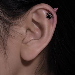 May include: A silver-toned cartilage earring featuring a black paw print design with a small, pointed spike backing. The jewelry is secured through the upper ear cartilage, showcasing a metallic finish and a distinct animal-inspired motif.