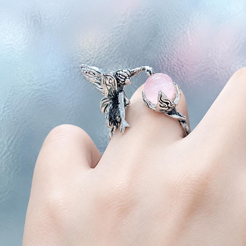 May include: An open-style ring crafted from oxidised silver-toned metal, featuring a detailed winged figure on one end and a smooth, round, pale pink stone held within a claw-like setting on the other end, creating a continuous wrap-around design.