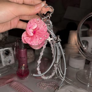 May include: A silver heart-shaped keychain featuring a large pink speckled orchid flower with small white pearls, a strand of translucent pink beads, a small white cross charm, and several thin silver metallic cords with small silver beads.