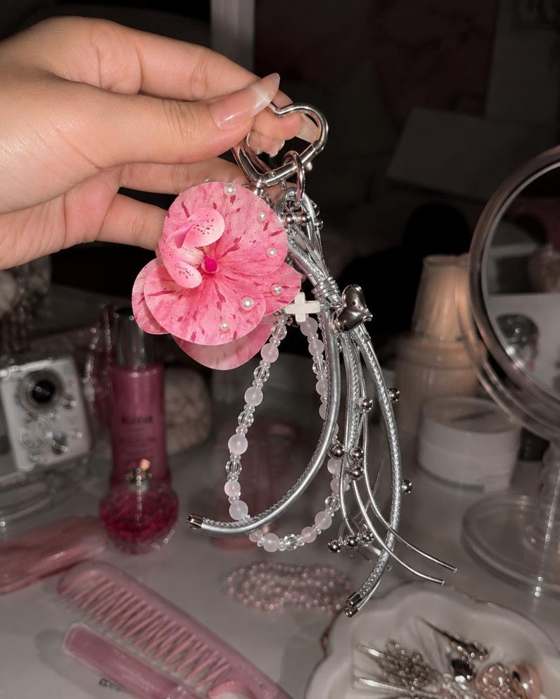 May include: A silver heart-shaped keychain featuring a large pink speckled orchid flower with small white pearls, a strand of translucent pink beads, a small white cross charm, and several thin silver metallic cords with small silver beads.