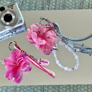 May include: Two decorative bag charms featuring pink floral elements. One charm includes a pink orchid, a silver heart-shaped clasp, and a light pink beaded strand. The other features a bright pink flower, a striped ribbon, and a small pink flip-flop charm.