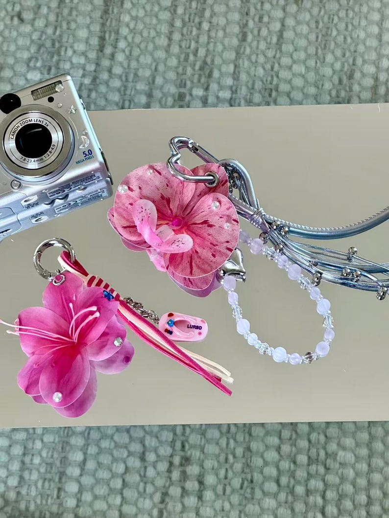 May include: Two decorative bag charms featuring pink floral elements. One charm includes a pink orchid, a silver heart-shaped clasp, and a light pink beaded strand. The other features a bright pink flower, a striped ribbon, and a small pink flip-flop charm.