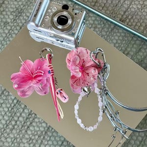 May include: A silver Canon PowerShot A95 digital camera positioned above two decorative bag charms. One charm features a pink flower, a pink cord, and a small flip-flop pendant. The other charm includes a pink orchid, a beaded chain, and silver ribbons.