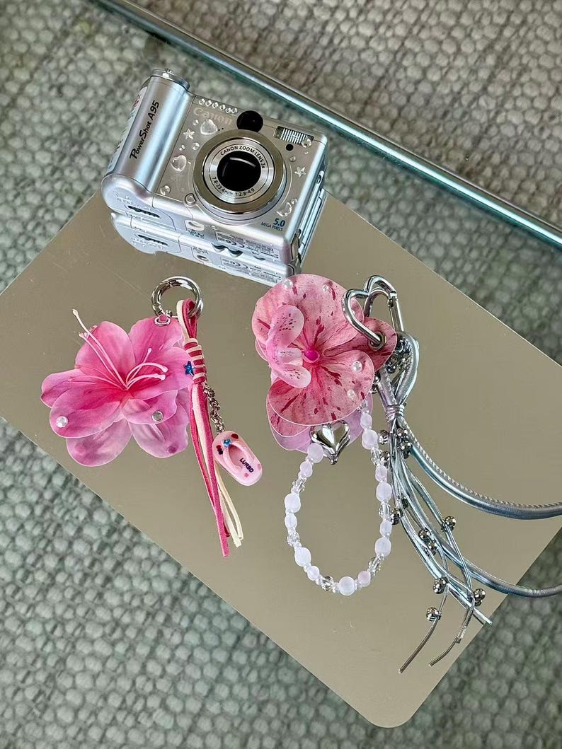 May include: A silver Canon PowerShot A95 digital camera positioned above two decorative bag charms. One charm features a pink flower, a pink cord, and a small flip-flop pendant. The other charm includes a pink orchid, a beaded chain, and silver ribbons.