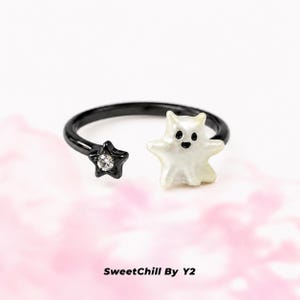 Puede incluir: Un anillo abierto negro con una figura de fantasma blanca en un extremo y una peque&ntilde;a estrella negra con un cristal transparente incrustado en el otro. El texto SweetChill By Y2 aparece debajo del anillo. El dise&ntilde;o es sencillo y contrastado.