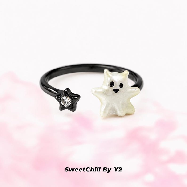 Puede incluir: Un anillo abierto negro con una figura de fantasma blanca en un extremo y una peque&ntilde;a estrella negra con un cristal transparente incrustado en el otro. El texto SweetChill By Y2 aparece debajo del anillo. El dise&ntilde;o es sencillo y contrastado.