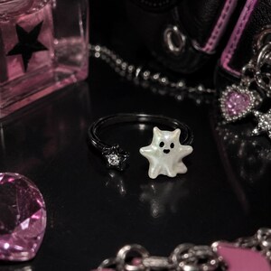 Puede incluir: Un anillo abierto negro que presenta un dije blanco nacarado con forma de fantasma en un extremo y un peque&ntilde;o cristal transparente con forma de estrella engastado en metal negro en el otro. El dije de fantasma tiene dos puntos negros por ojos.