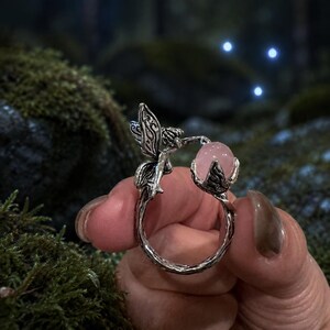 May include: A silver-toned metal ring featuring a textured band that resembles a twisted branch. One end of the ring depicts a small fairy with detailed wings, while the other end holds a smooth, round, pale pink gemstone in a claw setting.