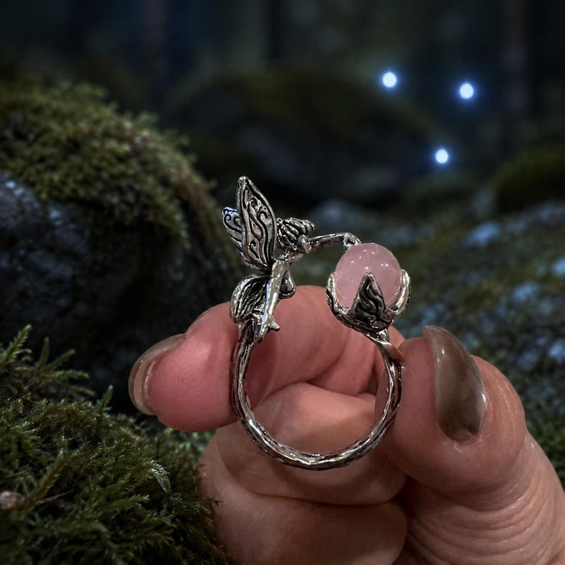 May include: A silver-toned metal ring featuring a textured band that resembles a twisted branch. One end of the ring depicts a small fairy with detailed wings, while the other end holds a smooth, round, pale pink gemstone in a claw setting.
