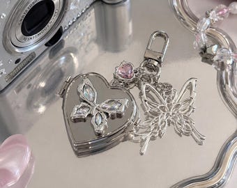 May include: A silver heart-shaped locket keychain featuring a central butterfly motif with iridescent crystals, a small pink crystal accent, and a dangling open-work silver butterfly charm. The text "SweetChill by Y2" is printed in orange.