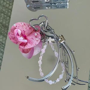 May include: A decorative keychain featuring a pink and white speckled orchid flower, a heart-shaped silver carabiner, a small white cross charm, a light pink beaded strand, and multiple silver-toned metallic ribbons with small spherical accents.