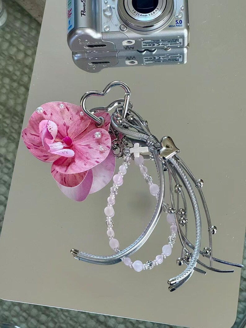 May include: A decorative keychain featuring a pink and white speckled orchid flower, a heart-shaped silver carabiner, a small white cross charm, a light pink beaded strand, and multiple silver-toned metallic ribbons with small spherical accents.