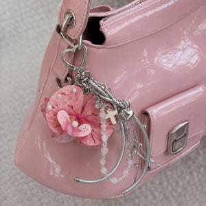 May include: A glossy light pink handbag featuring a silver-toned metal heart-shaped clasp. Attached to the hardware is a decorative charm set consisting of a pink orchid flower, a small white cross, and a strand of light pink beads on a silver chain.
