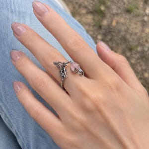 May include: A silver-toned open ring featuring a detailed fairy figure on one end and a small, round, pale pink gemstone held within a claw-like setting on the other end. The metal has an oxidised finish that highlights the intricate textures.