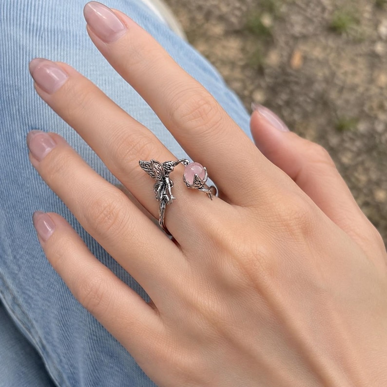 May include: A silver-toned open ring featuring a detailed fairy figure on one end and a small, round, pale pink gemstone held within a claw-like setting on the other end. The metal has an oxidised finish that highlights the intricate textures.