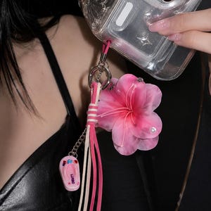 May include: A phone charm featuring a large, translucent pink flower with a small pearl accent, a miniature pink flip-flop charm with the text LURBO, and a multi-strand tassel in shades of pink and white, all attached to a silver metal ring.