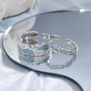 May include: Two silver-toned open rings featuring a textured, irregular metal surface with inlaid light blue enamel detailing. The rings have a reflective finish and are displayed on a mirror surface. The text Sweetchill By Y2 is visible.