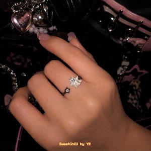 May include: An open-style ring featuring a small white ghost figure on one end and a black star-shaped charm with a clear center stone on the other end. The ring band is thin and dark in color, designed to be worn on a finger with an adjustable opening.