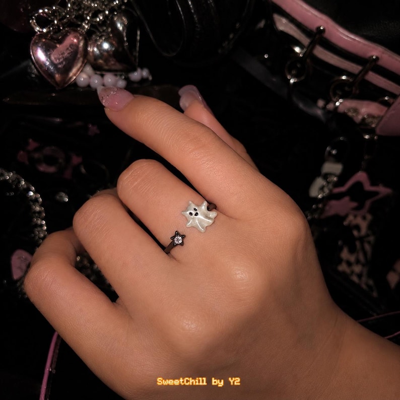 Puede incluir: Un anillo de estilo abierto que presenta una peque&ntilde;a figura de fantasma blanco en un extremo y un dije negro en forma de estrella con una piedra central transparente en el otro. La banda del anillo es delgada y oscura, dise&ntilde;ada con una abertura ajustable.