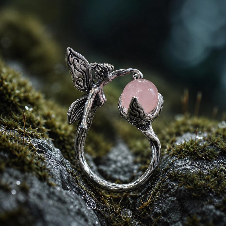 May include: A silver-toned ring featuring a fairy figure with detailed wings. The fairy holds a smooth, round, pale pink gemstone in a claw-like setting. The band of the ring has a textured, branch-like appearance, crafted from metallic material.