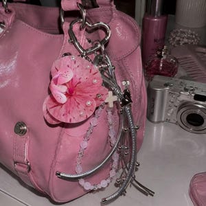 May include: A glossy pink handbag featuring a silver heart-shaped keychain attachment. The keychain includes a large pink floral charm with small white pearls, a string of light pink beads, a small white cross, and silver metallic accents.