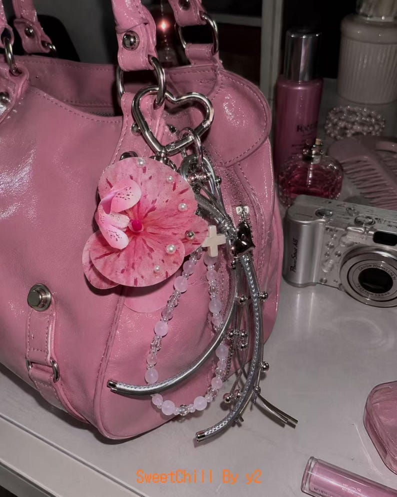 May include: A glossy pink handbag featuring a silver heart-shaped keychain attachment. The keychain includes a large pink floral charm with small white pearls, a string of light pink beads, a small white cross, and silver metallic accents.