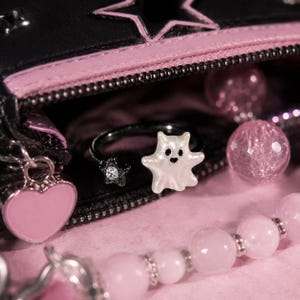 Puede incluir: Un anillo ajustable de metal negro con una figura de fantasma blanco en un extremo y una peque&ntilde;a estrella negra con un cristal transparente en el otro. El anillo est&aacute; colocado cerca de un estuche negro con cremallera, borde rosa y dije de coraz&oacute;n.