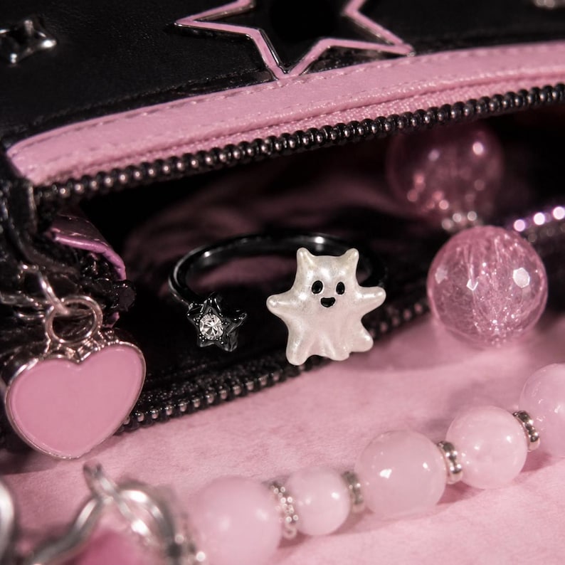 Puede incluir: Un anillo ajustable de metal negro con una figura de fantasma blanco en un extremo y una peque&ntilde;a estrella negra con un cristal transparente en el otro. El anillo est&aacute; colocado cerca de un estuche negro con cremallera, borde rosa y dije de coraz&oacute;n.