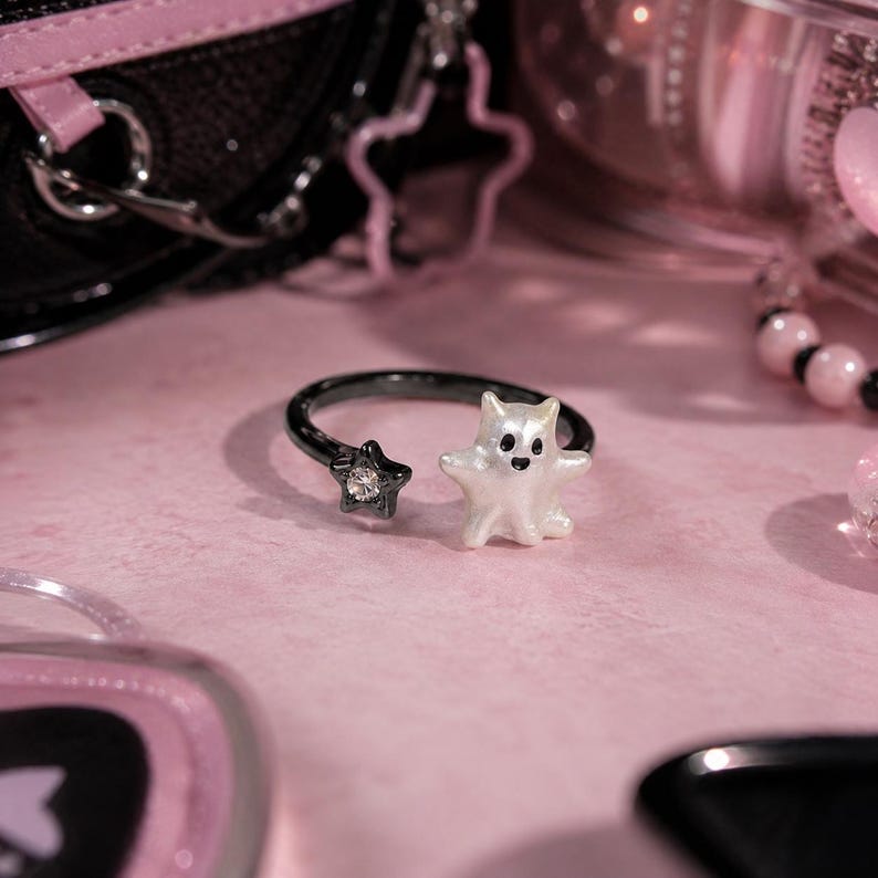 Puede incluir: Un anillo abierto de metal negro que presenta una peque&ntilde;a estrella negra con un estr&aacute;s transparente incrustado en un extremo y una figura de fantasma blanca nacarada con ojos negros y una peque&ntilde;a boca negra en el otro extremo.