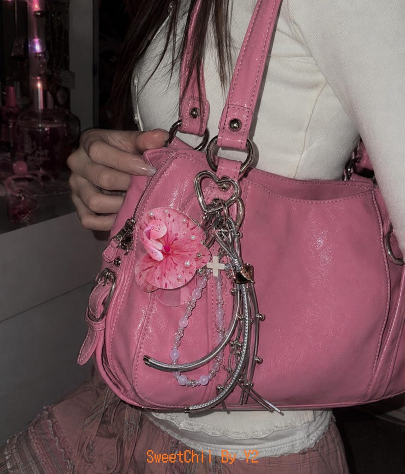 May include: A pink handbag with a glossy finish, featuring silver-toned hardware, dual shoulder straps, and a decorative cluster of charms including a pink flower, a white cross, and beaded strands. The text SweetChii by Y2 is visible at the bottom.