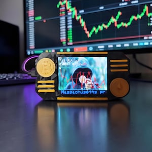 Op de afbeelding: Een zwart en geel cryptocurrency ticker-apparaat met een geometrisch getextureerde behuizing. Het scherm toont een rode lijngrafiek, de tekst BTCDOWN, 0,0012 $, -1,0% en Massachusetts pr. Een Bitcoin-logo staat links en een Ethereum-logo rechts.