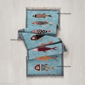 Peut inclure: Un chemin de table bleu clair orné d'un motif répétitif de poissons stylisés dans des tons marron, rouge et orange. Le tapis présente une bordure foncée avec un motif géométrique en losanges et des franges. Le texte HandKnotRugsIndia est visible.