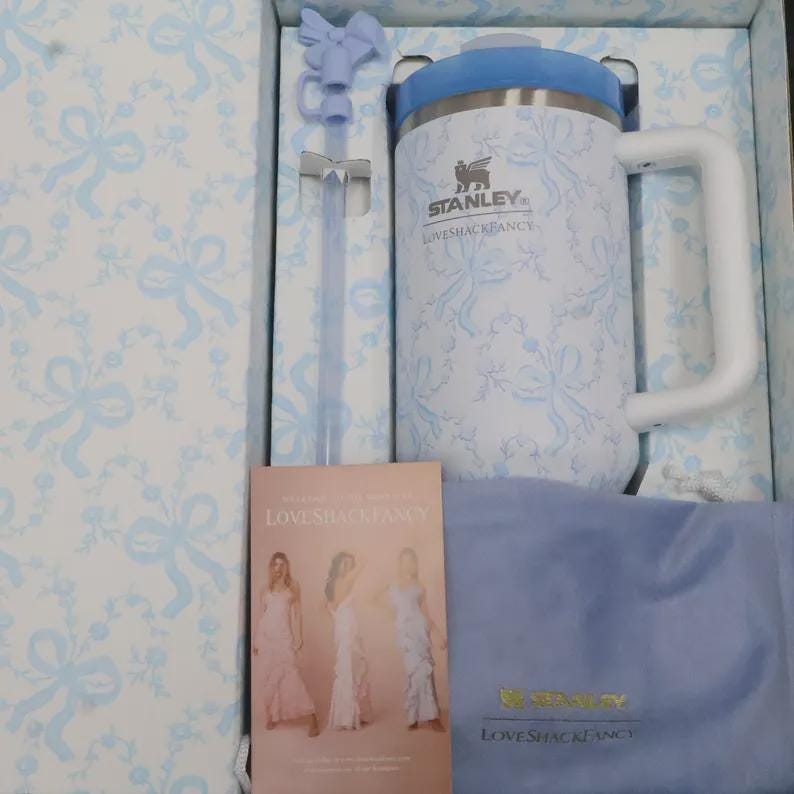 May include: A Stanley Loveshackfancy insulated mug with a white handle and blue lid, presented in a box with a matching bow pattern. Includes a straw, a soft blue cloth, and a Loveshackfancy card.