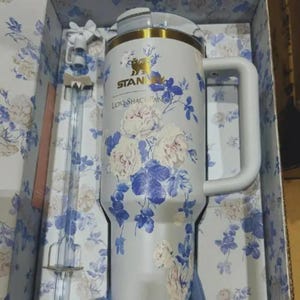 May include: A white insulated travel mug with a gold rim and handle, decorated with a floral pattern of white and blue flowers. The mug has the text "STAN" and "LOVESHACKFANCY". A matching straw and box are included.
