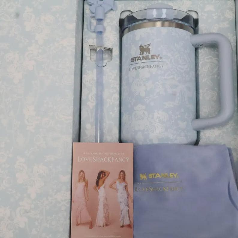 May include: A light blue Stanley Loveshackfancy mug with a handle, a straw, and a lid. The mug has a floral pattern and the Stanley logo. The set includes a small booklet and a blue cloth. The mug is packaged in a box with a floral design.