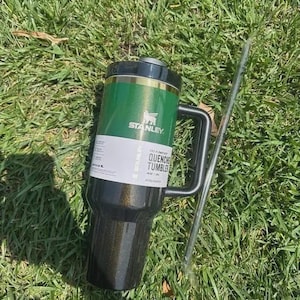 K&ouml;nnte beinhalten: Ein gr&uuml;ner und schwarzer Stanley Quencher-Becher mit schwarzem Griff und Deckel, der auf gr&uuml;nem Gras liegt. Der Becher hat ein wei&szlig;es Etikett mit dem Text "QUENCHER TUMBLER". Ein klarer Strohhalm liegt neben dem Becher.
