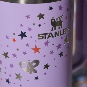 Może przedstawiać: Lawendowy kubek Stanley ze srebrną obręczą i uchwytem. Kubek posiada złote logo skrzydlatego niedźwiedzia i nazwę marki. Jest ozdobiony złotymi, pomarańczowymi i fioletowymi gwiazdami oraz srebrnym motywem motyla.