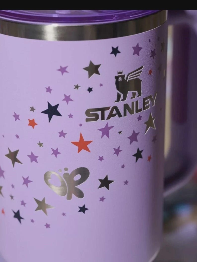 Może przedstawiać: Lawendowy kubek Stanley ze srebrną obręczą i uchwytem. Kubek posiada złote logo skrzydlatego niedźwiedzia i nazwę marki. Jest ozdobiony złotymi, pomarańczowymi i fioletowymi gwiazdami oraz srebrnym motywem motyla.