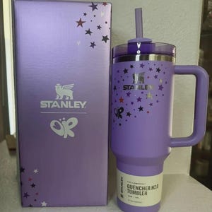Może przedstawiać: Fioletowy kubek Stanley Quencher H2.0 z uchwytem i słomką, obok pasującego pudełka. Zar&oacute;wno kubek, jak i pudełko mają wz&oacute;r gwiazd i logo Stanley. Pojemność: 1,18L.