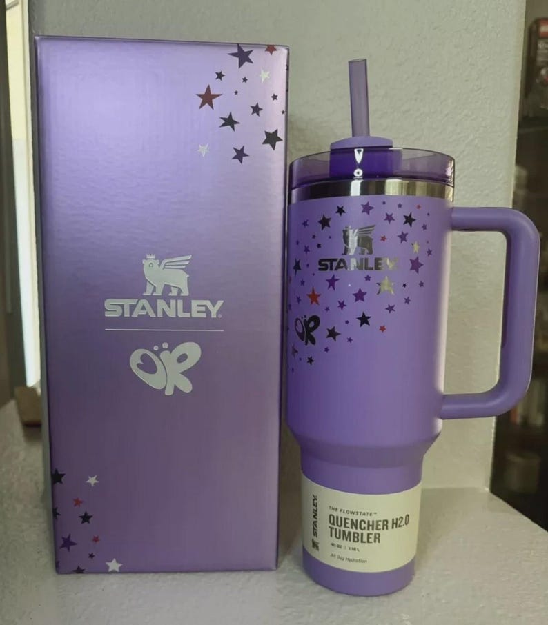 Może przedstawiać: Fioletowy kubek Stanley Quencher H2.0 z uchwytem i słomką, obok pasującego pudełka. Zar&oacute;wno kubek, jak i pudełko mają wz&oacute;r gwiazd i logo Stanley. Pojemność: 1,18L.