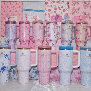 May include: A collection of Stanley tumblers in various pastel colors, adorned with floral patterns. Each tumbler features a handle, a lid, and a decorative figurine on top. The background showcases matching floral-patterned boxes.