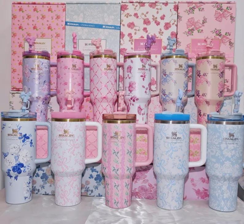 May include: A collection of Stanley tumblers in various pastel colors, adorned with floral patterns. Each tumbler features a handle, a lid, and a decorative figurine on top. The background showcases matching floral-patterned boxes.