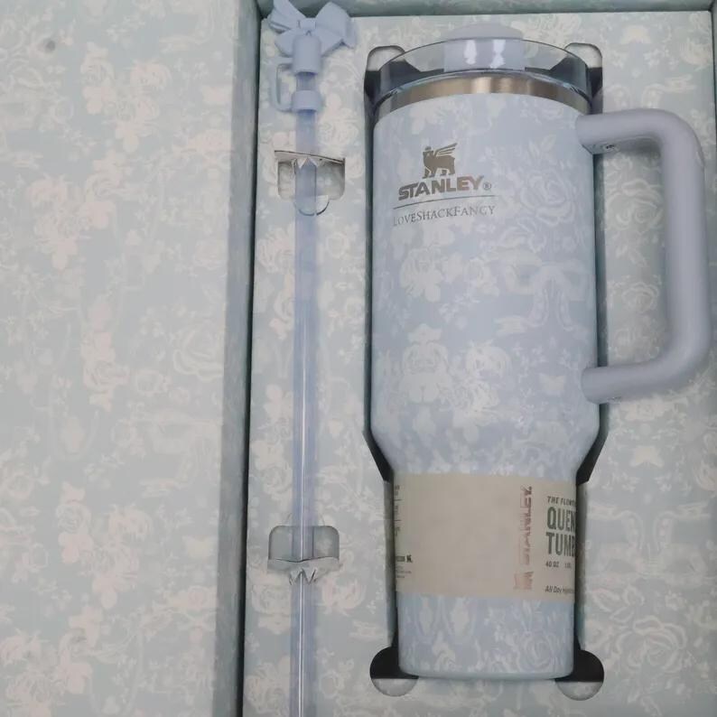 May include: A light blue Stanley Loveshackfancy floral print tumbler with a handle and a straw. The tumbler is in a matching floral print box. The tumbler has the text "THE FLORAL QUEEN TUMBLER" on the label.