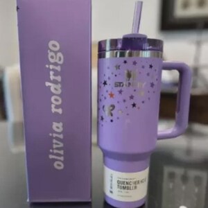 Może przedstawiać: Lawendowy kubek Stanley Quencher H2.0 FlowState z uchwytem, słomką i pokrywką. Kubek ma wz&oacute;r gwiazd i logo Stanley. Obok znajduje się pasujące pudełko z napisem "olivia rodrigo".