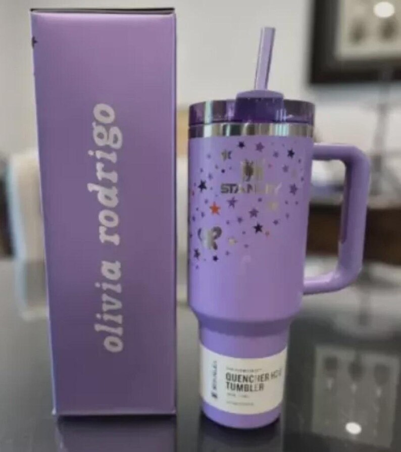 Może przedstawiać: Lawendowy kubek Stanley Quencher H2.0 FlowState z uchwytem, słomką i pokrywką. Kubek ma wz&oacute;r gwiazd i logo Stanley. Obok znajduje się pasujące pudełko z napisem "olivia rodrigo".