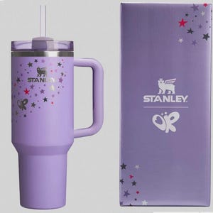 Może przedstawiać: Lawendowy kubek Stanley z uchwytem, słomką i pokrywką. Kubek jest ozdobiony srebrnymi gwiazdami i logo Stanley. Pasujące pudełko zawiera logo Stanley, gwiazdy i motyw motyla.