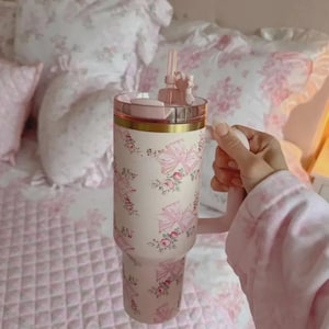 Pu&ograve; includere: Un grande bicchiere isolante con manico, con sfondo crema decorato con fiocchi rosa e motivi floreali. Il coperchio e il manico sono rosa, con una fascia dorata. Include cannuccia e decorazione.