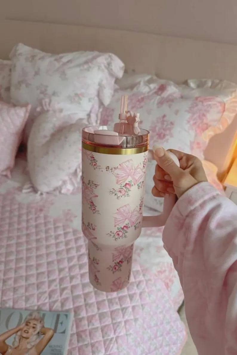 Pu&ograve; includere: Un grande bicchiere isolante con manico, con sfondo crema decorato con fiocchi rosa e motivi floreali. Il coperchio e il manico sono rosa, con una fascia dorata. Include cannuccia e decorazione.