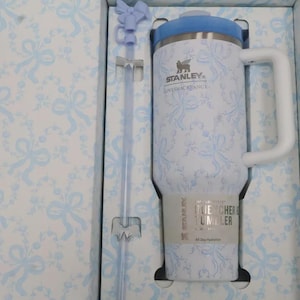 May include: A white and blue Stanley Quencher tumbler with a handle and straw, presented in a matching patterned box. The tumbler features a light blue lid and a floral bow design. The box and straw also have the same bow pattern.